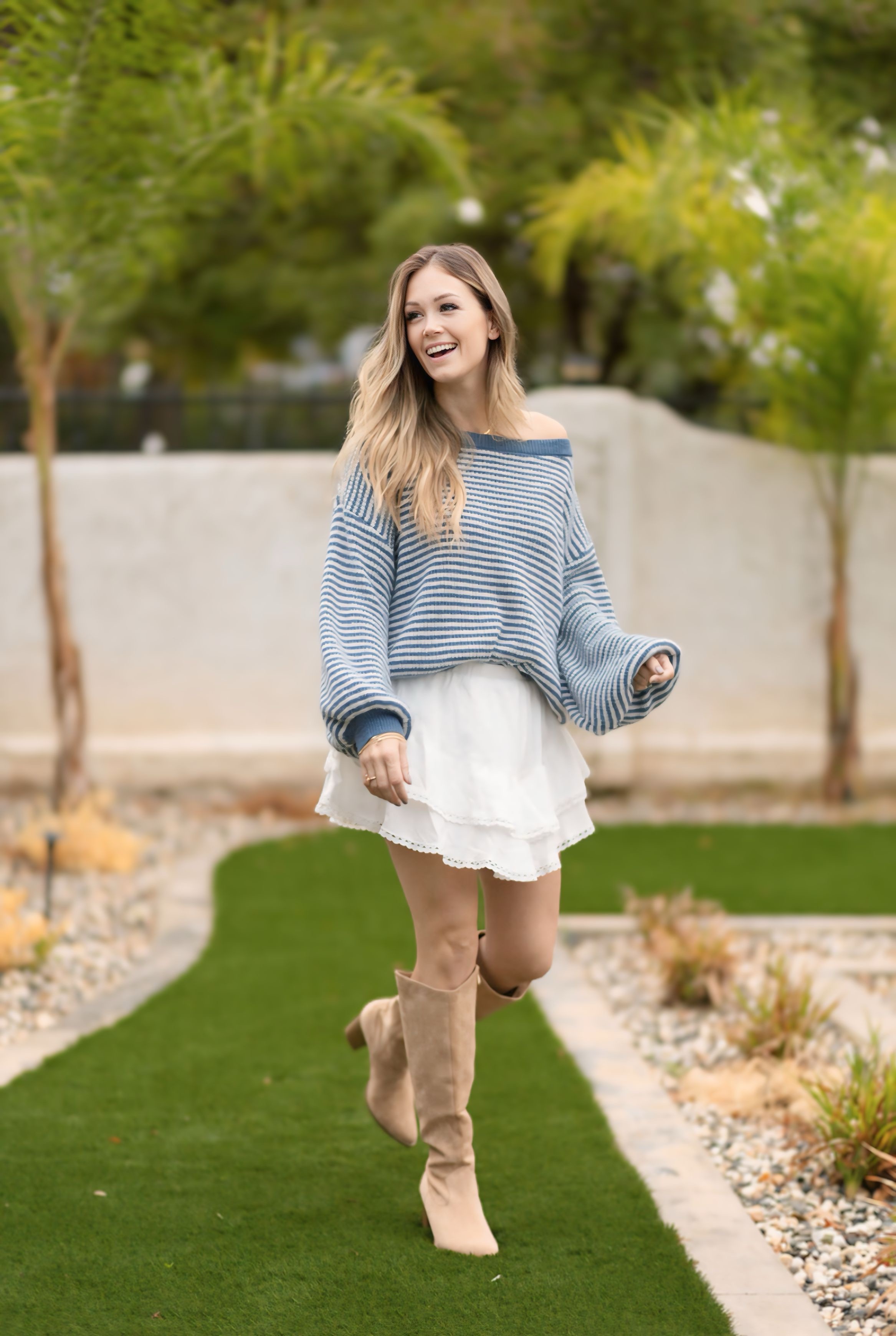 Elizabeth in a blue striped sweater and tan boots, smiling on a garden path.